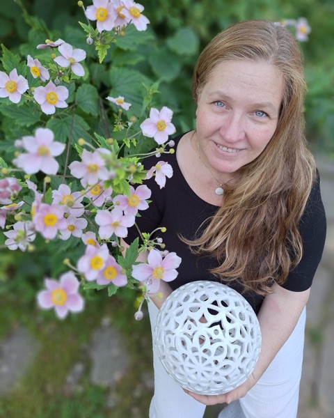 Steffi Gosch lächelt zwischen Blüten, in den Händen eine ihrer Lichtkugeln aus Keramik