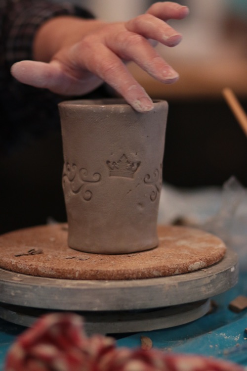 Hände drücken mit einem Stempel ein Muster in einen Becher aus Ton
